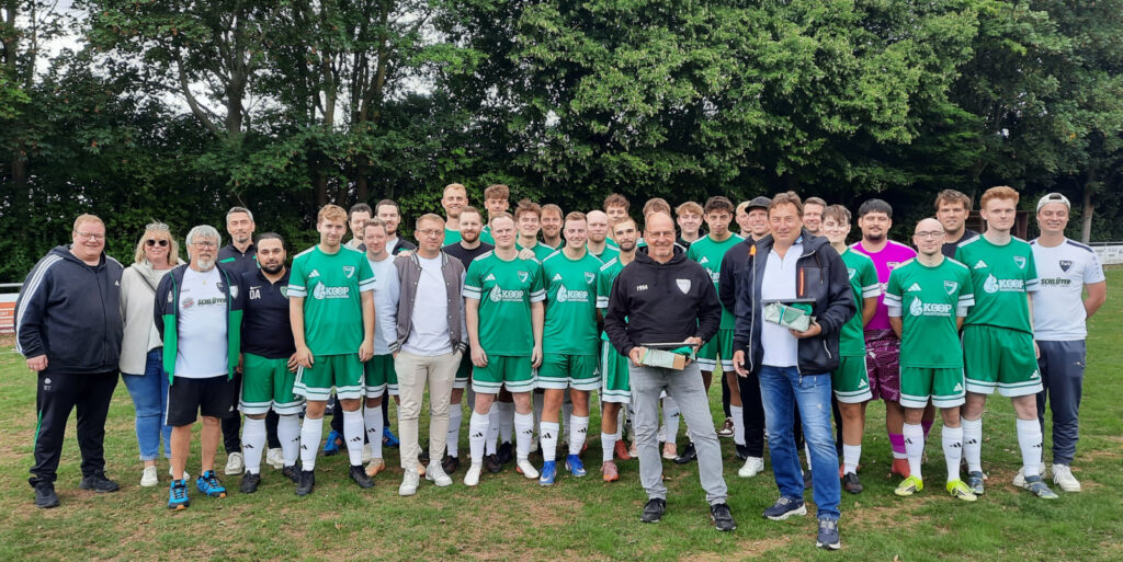 Eine Gruppe von Männern, die meisten davon in Fußballtrikots, stehen auf einem Fußballplatz.